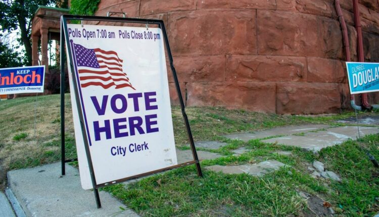 sign that reads "vote here, city clerk" with an american flag